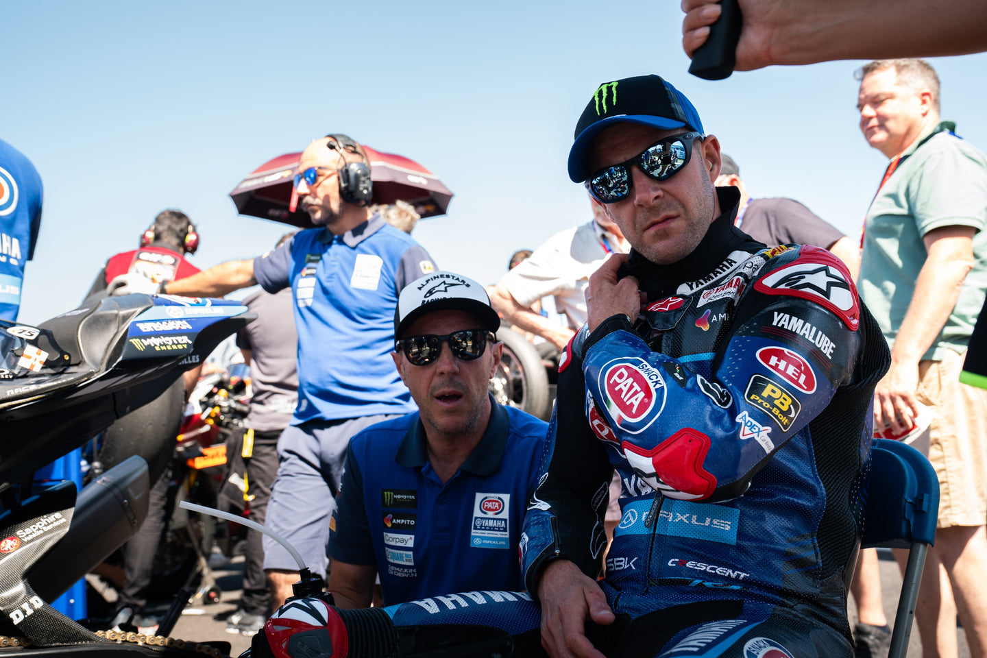 Jonathan Rea in a colorful Alpinestars suit with sponsor logos sitting on a motorcycle, surrounded by people and racing equipment.